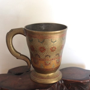 May include: A brass mug featuring a curved handle and a pedestal base. The exterior surface is decorated with intricate etched patterns in red, gold, and dark tones, arranged in horizontal bands around the body of the vessel.