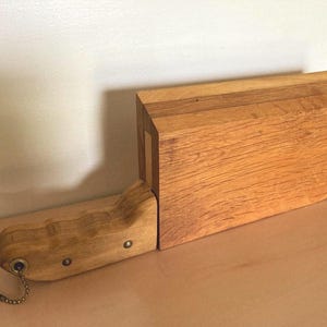 May include: A wooden knife block with a matching handle. The block is rectangular with a curved front edge. The handle is attached with small metal rivets and a chain. The wood has a natural, light brown color.