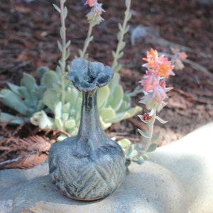 May include: A small, hand-formed ceramic bud vase with a dark, mottled blue-grey glaze. The vase features a narrow neck that flares into a ruffled, flower-like opening at the top and a rounded, textured base with a subtle cross-hatched pattern.