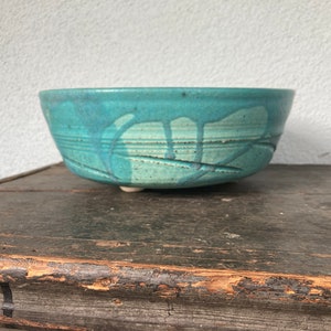 May include: A round ceramic bowl with a teal and turquoise glaze featuring a drip pattern and horizontal incised lines around the exterior. The bowl has a wide opening and a slightly tapered base, showcasing a speckled texture throughout the surface.