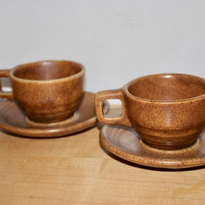 May include: Two brown ceramic coffee cups with matching saucers. The cups have square handles and a speckled texture. The saucers are round with a slightly raised edge. The set is a warm, earthy tone, suitable for serving coffee or tea.