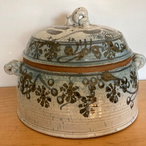 May include: A ceramic lidded serving dish with a light blue and brown floral design. The dish has a round shape with handles and a decorative knob on the lid. The design includes floral and vine patterns.