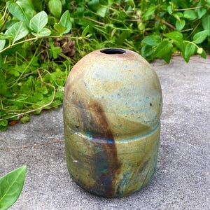 May include: A cylindrical ceramic vase with a rounded top and a narrow opening. The surface features a mottled glaze in shades of olive green, brown, and blue, with a distinct horizontal groove encircling the middle of the vessel's body.