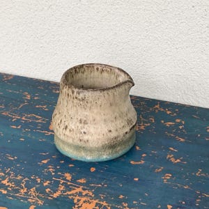 May include: A small ceramic creamer pitcher with a rounded body and a small spout. The exterior features a speckled cream and beige glaze on the upper portion, transitioning into a darker brown and vibrant blue band at the base of the vessel.