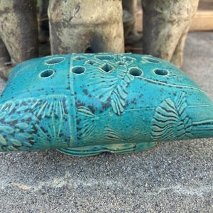 May include: A turquoise ceramic flower frog with a rectangular shape and multiple holes for arranging flowers. The surface features embossed botanical designs and a textured finish. This decorative piece is ideal for floral arrangements.