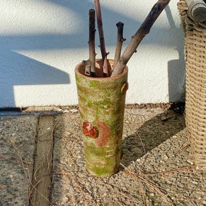 May include: A cylindrical, handmade ceramic vase with a textured, mossy green and brown finish. The vase contains several brown twigs. A woven basket is visible in the background.