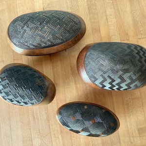 May include: Four oval-shaped wooden boxes with smooth, rounded edges. Each box features a distinct dark, intricate geometric pattern etched into the lid, contrasting with the natural light brown wood grain of the base and sides.
