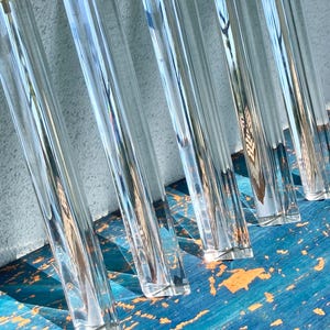 May include: Five clear, transparent, elongated acrylic rods with a triangular cross-section. Each rod features smooth, flat surfaces that catch and refract light, creating sharp, linear reflections along their vertical length.