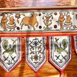 May include: A cream-colored fabric wall hanging featuring intricate embroidery. The top horizontal panel displays figures, a cow, and floral motifs. Below, four pointed pennants hang, each decorated with green bird illustrations and floral patterns, finished with red trim and tassels.