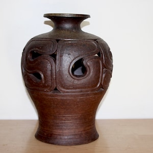 May include: A dark brown ceramic vase with a textured, matte finish. The upper body features large, open, curved cutouts arranged in a repeating pattern, while the lower base is solid with horizontal incised lines circling the circumference.