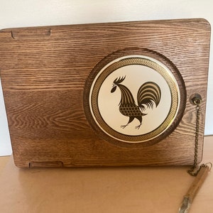 May include: A vintage wooden serving tray with a circular, decorative insert featuring a brown rooster design on a cream background. The tray has a rectangular shape with a handle and a metal chain attached to a small wooden utensil.