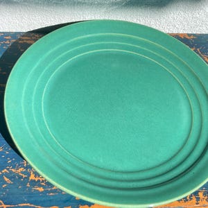 May include: A teal-colored oval plate with concentric ridges. The plate has a matte finish and is set against a blue and orange distressed wooden surface. The plate's design is simple and elegant.