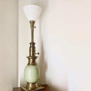May include: A vintage floor lamp with a white conical shade, brass stem, and a light green, oval-shaped base. The lamp has a square brass base and stands approximately 60 inches tall. It is a classic piece of home decor.
