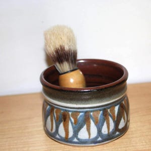 May include: A ceramic shaving brush holder with a decorative pattern in blue, brown, and white. The holder contains a shaving brush with a wooden handle and light brown bristles. The holder is on a wooden surface.