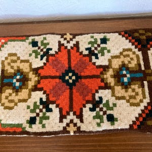 May include: A rectangular needlepoint runner featuring a central orange geometric floral motif on an off-white background. The design includes symmetrical patterns in brown, green, blue, and red, finished with yellow fringe on the left edge.
