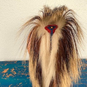 May include: A unique, handcrafted hair accessory featuring a mix of brown, tan, and white fur. The design includes a central decorative element with red, blue, and gold accents. The accessory is likely for ceremonial or decorative purposes.