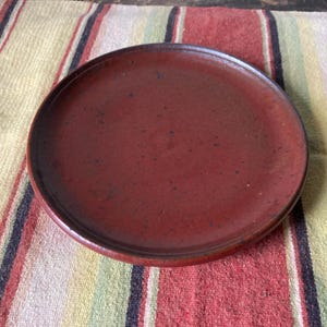 May include: A round, reddish-brown ceramic plate with a slightly raised edge. The plate has a matte finish and sits on a striped cloth with red, yellow, and black stripes. The plate is designed for serving food.