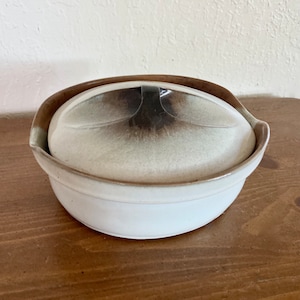 May include: A ceramic serving bowl with a lid. The bowl is white with a brown rim and a matching lid. The lid has a unique shape with a dark brown center. The bowl is sitting on a wooden surface.