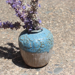 May include: A small, handmade ceramic vase with a light blue glazed top and a textured, unglazed bottom. The vase holds a bundle of dried lavender flowers. The vase is approximately 4 inches tall.