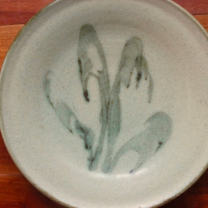 May include: A handmade ceramic plate with a speckled, off-white glaze. The plate features a stylized, abstract floral design in shades of gray-green. The rim has a darker green edge. The plate is set on a wooden surface.