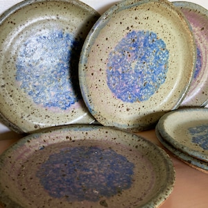 May include: A collection of round ceramic plates featuring a speckled beige base with a central blue and purple textured pattern. The plates have a raised rim and a glazed finish, with some stacked and others standing upright in a group.