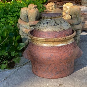 May include: A handmade, rustic ceramic pot with a lid. The pot is primarily reddish-brown with a speckled tan and brown lid. The pot has a wide, rounded body with a textured surface and a small spout. The pot is surrounded by a group of stone figures.