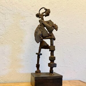 May include: A sculpture constructed from dark brown rusted metal hardware components including bolts, nuts, and washers. The figure stands on a rectangular wooden base, featuring an abstract, jagged torso and a circular head element made of metal.