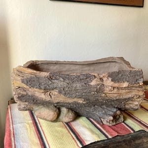 May include: A rectangular planter designed to resemble a weathered log. The planter is primarily brown with textured bark details and a naturalistic appearance. Two small stones are attached to the base. Ideal for rustic decor.