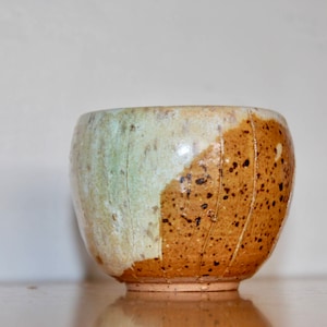 May include: Handmade ceramic bowl with a unique design. The bowl features a two-tone color scheme, with a light green and off-white section on the left and a speckled brown section on the right. The bowl has a rounded shape and a textured surface.
