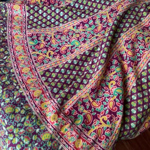 May include: A fabric textile featuring a deep burgundy base color with a repeating pattern of small green and yellow floral motifs. A wide border displays a larger, intricate design of yellow paisley shapes and green leaves on a red background.
