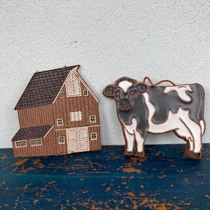May include: A rustic wooden barn and a cow-shaped ornament. The barn is brown with a dark roof and white accents. The cow is black and white with a brown outline. Both are placed on a dark blue surface.