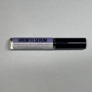 Op de afbeelding: Een heldere glazen tube Growth Serum met een zwarte dop. Het label is lavendelkleurig met witte tekst met de tekst "Growth Serum" en "Promotes Eyelash & Eyebrow Growth". De tube is 24,6 ml.