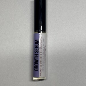 Op de afbeelding: Een cilindrische fles Growth Serum voor wimpers en wenkbrauwen. De fles heeft een zwarte dop en een heldere glazen body met een label met de tekst "GROWTH SERUM" en details over de biologische en veganistische samenstelling van het product.