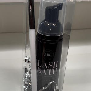 Può includere: Un flacone nero per il bagno delle ciglia con tappo trasparente e un pennello per trucco bianco e nero, confezionato in una scatola di plastica trasparente. Il flacone riporta la scritta "JLuxe LASH BATH". La scatola contiene coriandoli metallici argentati.