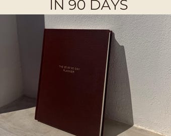 May include: A maroon-colored planner titled "The 30 60 90 Day Planner" is propped up against a wall. The text "Transform Your Life In 90 Days" is at the top of the image. The planner has a simple, elegant design.