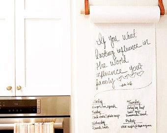 May include: A white wall mounted roll of paper with a wooden dowel and leather straps. The paper has handwritten text that reads "If you want lasting influence in the world, influence your family - Eds Witt". Below the text is a handwritten weekly meal plan.