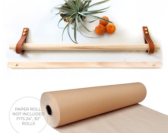 May include: Wooden paper towel holder with brown leather straps and gold metal accents. The holder is mounted on a white wall. A roll of brown paper is shown below the holder. The text on the paper roll reads "Paper Roll Not Included Fits 24", 30" Rolls".