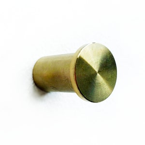 Op de afbeelding: Een gouden metalen haak met een ronde, platte bovenkant. De haak is bevestigd aan een witte muur.