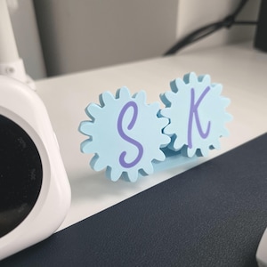 K&ouml;nnte beinhalten: Ein hellblaues Schreibtischornament bestehend aus zwei ineinandergreifenden zahnradf&ouml;rmigen Teilen. Jedes Zahnrad hat einen violetten Buchstaben in der Mitte aufgedruckt, wobei der Buchstabe S auf dem linken und der Buchstabe K auf dem rechten Zahnrad steht.