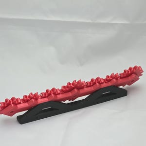 K&ouml;nnte beinhalten: Ein langes, rotes, gelenkiges, zahnradartiges Objekt mit einer Reihe verbundener, zahnf&ouml;rmiger Segmente, das horizontal auf einem schwarzen, dreieckigen St&auml;nder montiert ist. Das rote Bauteil weist ein wiederkehrendes Muster kleiner Spitzen auf.