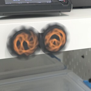 K&ouml;nnte beinhalten: Zwei kreisf&ouml;rmige schwarze Kn&ouml;pfe mit orangefarbenen Spiralmustern drehen sich schnell und erzeugen einen Bewegungsunsch&auml;rfe-Effekt. Die Kn&ouml;pfe sind auf einer wei&szlig;en horizontalen Fl&auml;che unter einem schwarzen elektronischen Bedienfeld montiert.