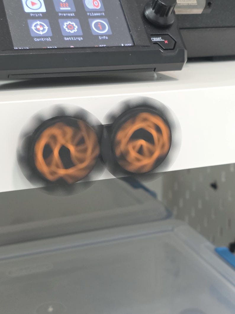 K&ouml;nnte beinhalten: Zwei kreisf&ouml;rmige schwarze Kn&ouml;pfe mit orangefarbenen Spiralmustern drehen sich schnell und erzeugen einen Bewegungsunsch&auml;rfe-Effekt. Die Kn&ouml;pfe sind auf einer wei&szlig;en horizontalen Fl&auml;che unter einem schwarzen elektronischen Bedienfeld montiert.