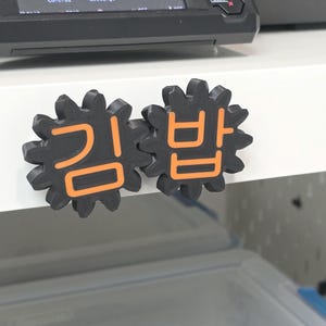 K&ouml;nnte beinhalten: Zwei schwarze, zahnradf&ouml;rmige Kunststoffobjekte mit aufgedruckten orangefarbenen koreanischen Schriftzeichen auf der Vorderseite. Die Zeichen lauten "김밥". Diese Gegenst&auml;nde sind an einer wei&szlig;en Oberfl&auml;che unter einem 3D-Drucker-Display befestigt.