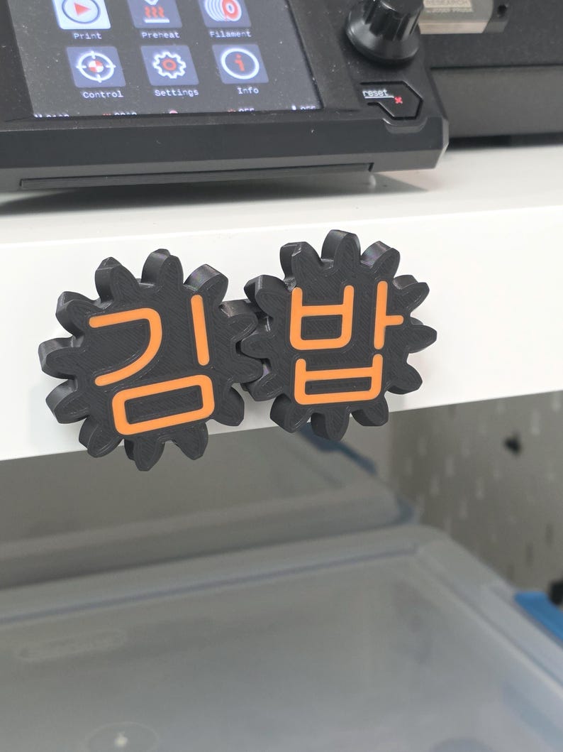 K&ouml;nnte beinhalten: Zwei schwarze, zahnradf&ouml;rmige Kunststoffobjekte mit aufgedruckten orangefarbenen koreanischen Schriftzeichen auf der Vorderseite. Die Zeichen lauten "김밥". Diese Gegenst&auml;nde sind an einer wei&szlig;en Oberfl&auml;che unter einem 3D-Drucker-Display befestigt.