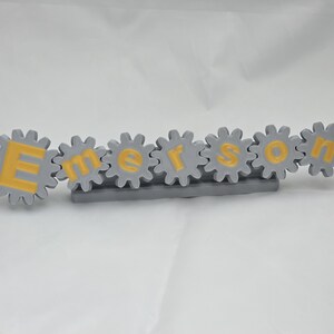 K&ouml;nnte beinhalten: Ein graues Namensschild in Zahnradform mit dem Namen Emerson in gelben Gro&szlig;buchstaben. Jeder Buchstabe ist in einem einzelnen grauen Zahnrad zentriert, und die gesamte Anordnung ist auf einer rechteckigen grauen Basis montiert.