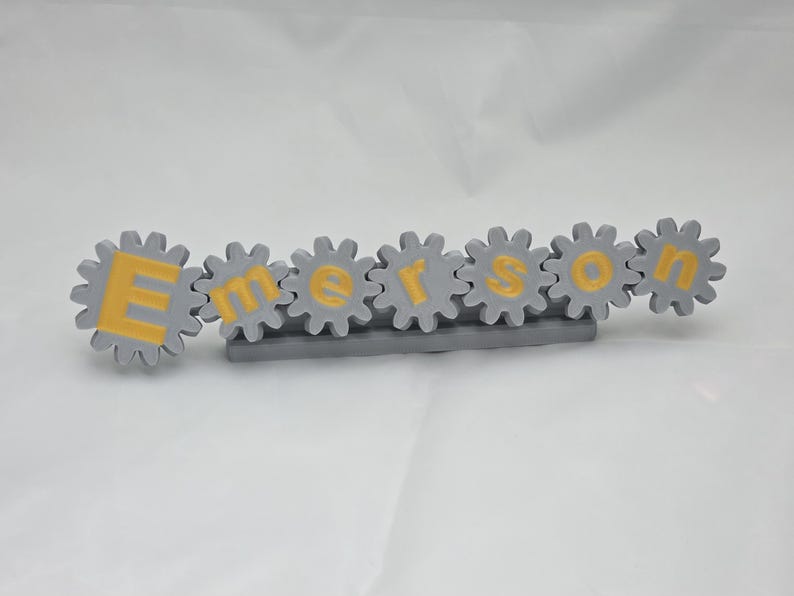 K&ouml;nnte beinhalten: Ein graues Namensschild in Zahnradform mit dem Namen Emerson in gelben Gro&szlig;buchstaben. Jeder Buchstabe ist in einem einzelnen grauen Zahnrad zentriert, und die gesamte Anordnung ist auf einer rechteckigen grauen Basis montiert.