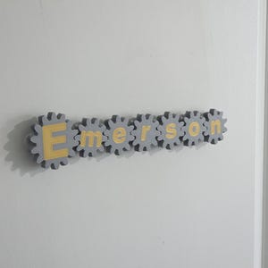 K&ouml;nnte beinhalten: Ein an der Wand montiertes Namensschild mit der Aufschrift Emerson, bestehend aus sieben einzelnen grauen zahnradf&ouml;rmigen Teilen. Jedes Zahnrad enth&auml;lt einen einzelnen gelben Buchstaben in der Mitte, was eine mechanische Optik erzeugt.