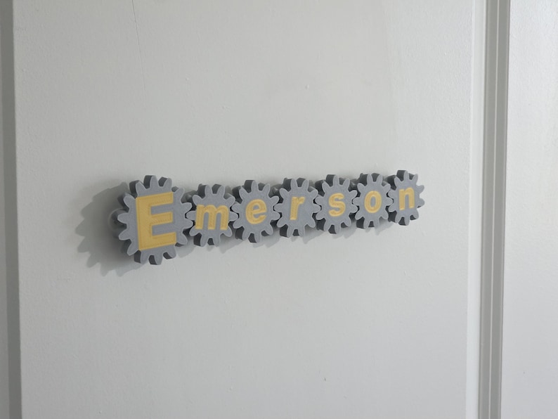 K&ouml;nnte beinhalten: Ein an der Wand montiertes Namensschild mit der Aufschrift Emerson, bestehend aus sieben einzelnen grauen zahnradf&ouml;rmigen Teilen. Jedes Zahnrad enth&auml;lt einen einzelnen gelben Buchstaben in der Mitte, was eine mechanische Optik erzeugt.