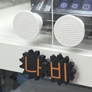 K&ouml;nnte beinhalten: Ein 3D-Drucker-Geh&auml;use mit zwei wei&szlig;en, runden L&uuml;ftungsgittern und zwei schwarzen, zahnradf&ouml;rmigen Kn&ouml;pfen mit orangefarbenen koreanischen Schriftzeichen. Das Display des Druckers zeigt den Text Original Prusa MK4S.