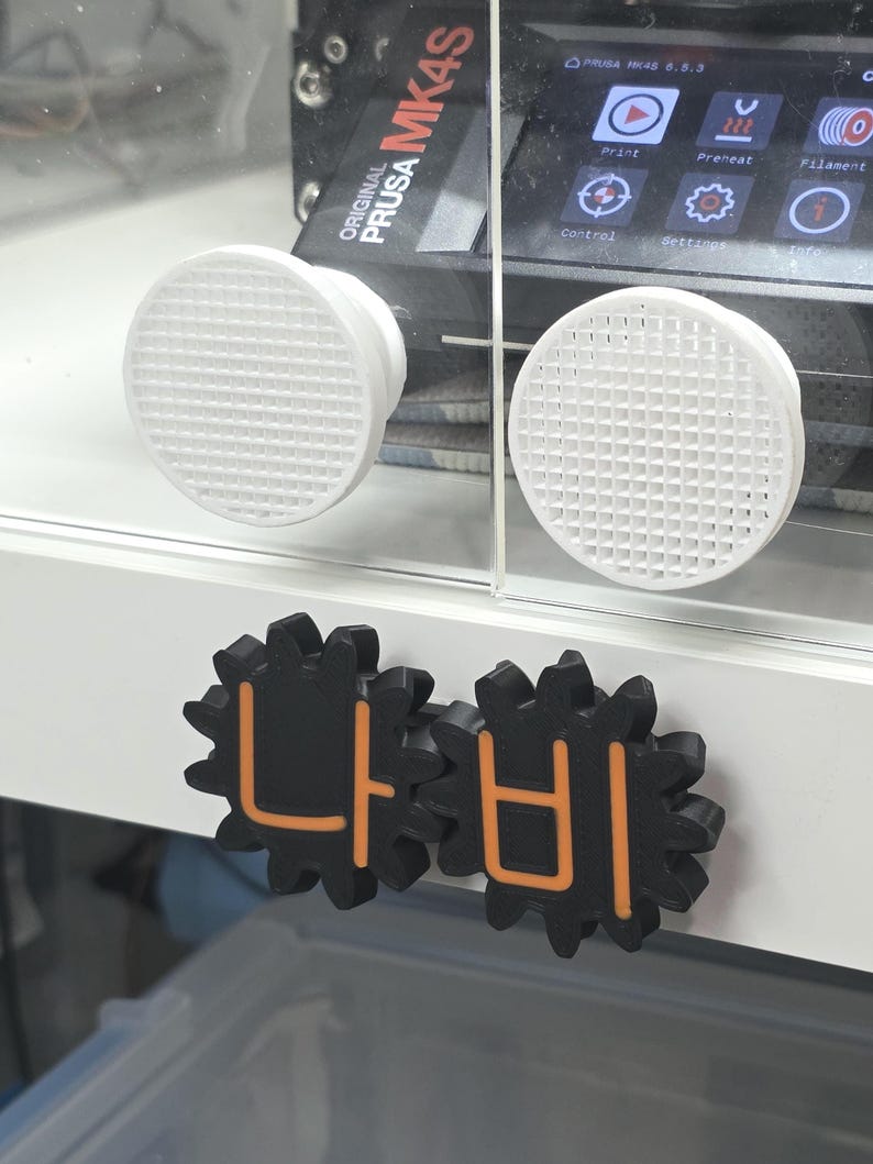 K&ouml;nnte beinhalten: Ein 3D-Drucker-Geh&auml;use mit zwei wei&szlig;en, runden L&uuml;ftungsgittern und zwei schwarzen, zahnradf&ouml;rmigen Kn&ouml;pfen mit orangefarbenen koreanischen Schriftzeichen. Das Display des Druckers zeigt den Text Original Prusa MK4S.